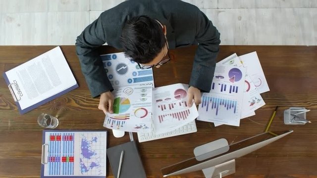 Top view shot of businessman in suit and glasses sitting at table and going through financial documents. He is comparing data and typing on computer