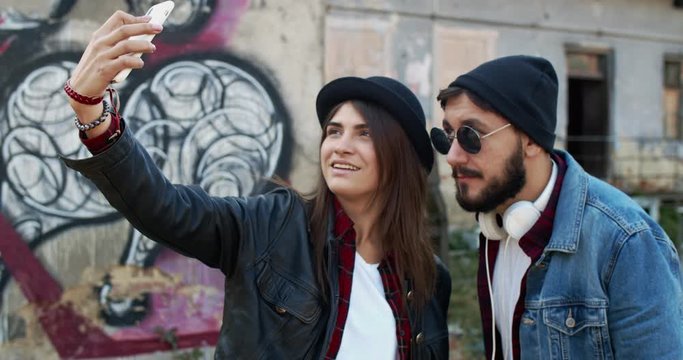 Nice selfie photos on the smartphone camera being taken by joyful young happy hipsters boyfriend and girlfriend outdoor in the slums at the graffity on the wall. Close up.