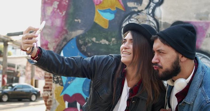 Cheerful young Caucasian stylish girl and guy, friends hipsters, laughing and posing funny to the smartphone camera while taking selfie photos. Outdoors in sunlight at the graffity on the wal.
