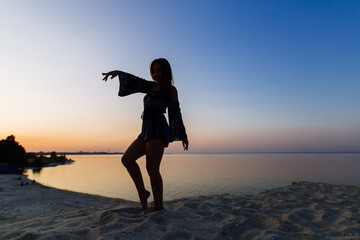 Fototapeta premium silhouette of a slim woman on sea beach which doing fitness and warm-up with copy space for text. Young loneliness woman enjoys sunset on a sandy beach on blue sky background