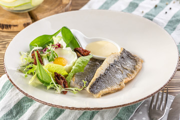 Fried white fish fillet with fried egg and greens on white plate on wooden table