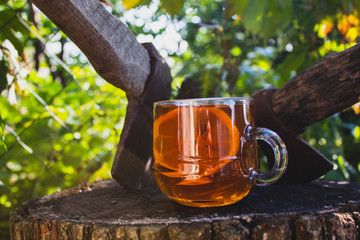 A transparent cup with tea and lemon stands near two axes. On a natural background. Close-up.