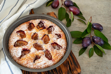 Shaped plum pie, dishcloth, plum branches and fruits on a light wooden background.