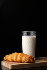 A glass with milk and one french croissant on a wooden board. Black background. White drink. Cow's milk. Classic french croissant. Homemade baking.
