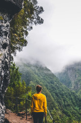 Young woman in yellow sweatshirt on viewpoint in Levada Caldeirao Verde, Madeira, Portugal. Green scenic mountains in mist, foggy landscape. Female traveler. Instagram, hipster filter. Vertical photo