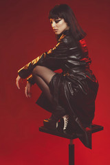 Attractive young woman in black leather coat and stockings on black shoes alluring posing. Fashion shoot in red studio. Sexy poster with pretty brunette girl. female sitting on chair