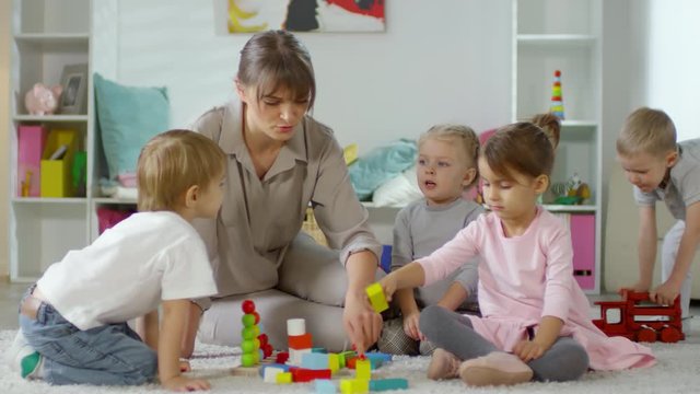 Group of little girls and boys and female teacher sitting together on the floor in kindergarten room, talking and playing with educational toys
