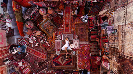 Beautiful happy girl in a long white dress laying on the carpet and rugs in Goreme, Cappadocia, Turkey. Top view drone.