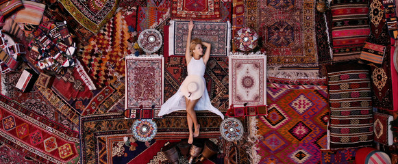 Beautiful happy girl in a long white dress laying on the carpet and rugs in Goreme, Cappadocia, Turkey. Top view drone.