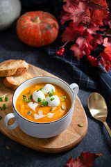 Pumpkin soup with seeds, mozzarella balls and greenery in white bowl on dark background with autumn leaves and pumpkins. Close up. Menu, restaurant. Vegeterian seasonal food.
