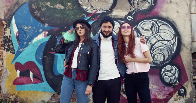 Portrait of the young stylish Caucasian friends hipsters at the graffity on the wall outside, man and two women looking and posing to the camera.