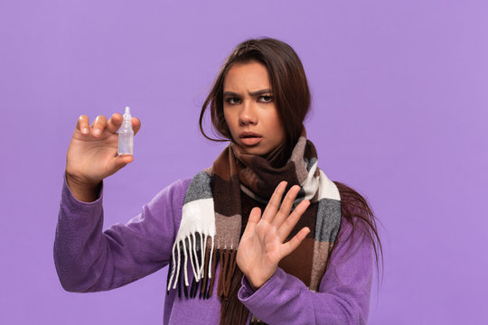 Pretty african-american girl in a sweater and cosy checked scarf using nasal spray receive treatment for running nose standing isolated over purple background. Catching cold in winter.
