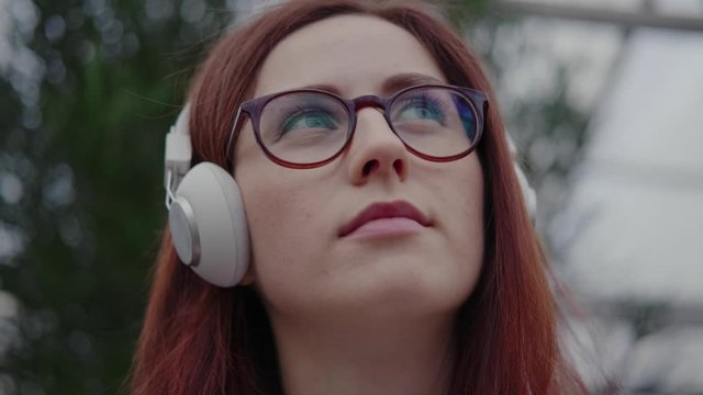 Portrait of a modern young woman with red hair, in big headphones and glasses: she listens to music and looks up, smiles easily. Concept of modern generation, young adult. Close up