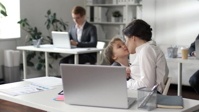 Beautiful businesswoman hugging and kissing her little son at office desk during workday while her male colleagues working in the background