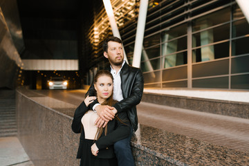 The guy hugs his girlfriend, followed by a car in the distance with the lights on. The couple looks into the future. Stylish couple in love with strict clothes black and white with beige