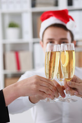 A group of businesspeople celebrating Christmas New Year corporate in the office while toasting holding glasses of champagne in hands on their heads wearing Santa cap.