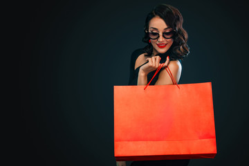Fototapeta na wymiar Black Friday sale concept for shop. Shopping woman in sunglasses holding red bag isolated on dark background.
