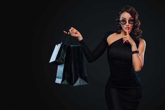 Black Friday sale concept for shop. Girl in sunglasses holding big bag isolated on dark background at shopping. Woman pointing to looking on copy space for sales text.