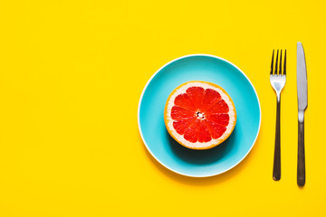 Concept of vegan diet. High angle shot - Appetizing juicy fresh grapefruit on color plate with fork and knife on yellow background with copy space.