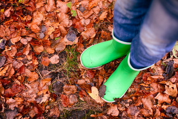 Child in green rubber boots standing on fallen leaves.