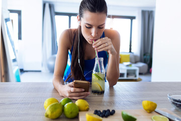 Beautiful sporty young woman drinking detox juice while listening to music with smartphone in the kitchen.