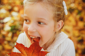 Happy little girl playing with autumn leaves. ute funny girl with open mouth in autumn park.Close up portrait of lovely girl with emotional face fooling and making faces. Smiling and laughing kid.