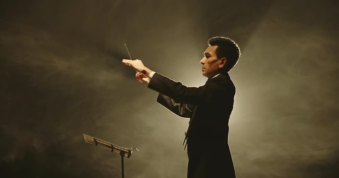 Male orchestra conductor controlling music in orchestra pit by movement of his hands and white baton, studio shot on black background 4k footage