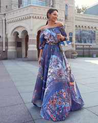 Young beautiful brunette woman with long straight hair in a tail wearing colorful blue dress, holding gray leather handbag and walking old city street