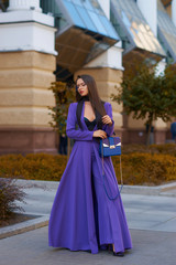 Young beautiful caucasian woman with long straight hair and makeup in purple trousers and long coat cape standing and posing at city street in autumn