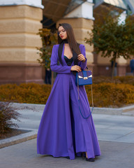 Young beautiful caucasian woman with long straight hair and makeup in purple trousers and long coat cape standing and posing at city street in autumn