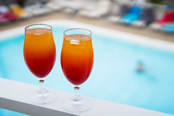 Two glass glasses with a red and yellow cocktail stand on a white narrow surface. in the background there is a swimming pool and beach umbrellas. Blurred background. Selective focus. Copy space.