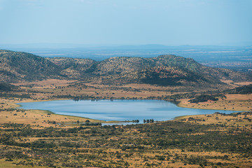 A dam/lake at the foot of the hills, surrounded by the dry and bushy plains of Pilanesberg Nature Reserve in South Africa
