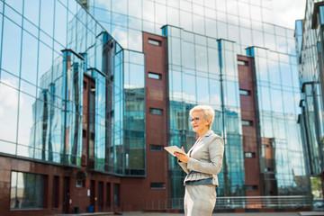 stylish adult woman in age in a suit and glasses against the background of a glass building with a tablet in her hands