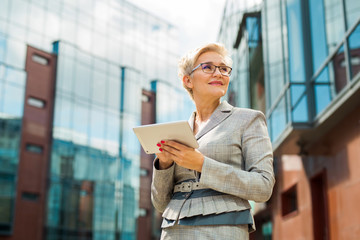 stylish adult woman in age in a suit and glasses against the background of a glass building with a tablet in her hands