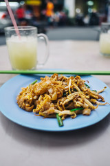 3/4 view Malaysian pad thai rice noodle dish with shrimp and green onion served on blue plate alongside citrus juice. Hawker fare street food served outdoors with street scene visible in background.