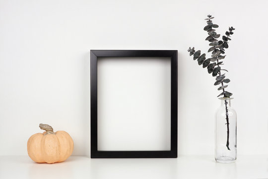 Mock up black frame with pumpkin decor and eucalyptus branch on a shelf or desk. Autumn concept. Portrait frame against a white wall.