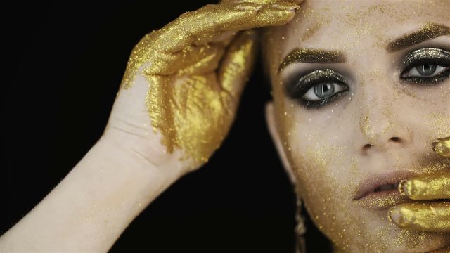 Girl face with golden make-up  in the studio on  black background.  Gold paint on the face and fingers. Hand covers face Gold earrings, gold eye shadow, clear skin. 