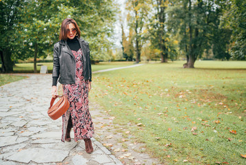 Smiling female dressed boho fashion style colorful long dress with black leather biker jacket with brown leather flap bag having a autumn park walking.