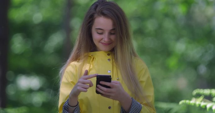  Young Smiling Woman with Mobile phone texting in a Forest. Girl in a Yellow Parka in the Woods. Pretty Hiker with Cell sending a message within tree foliage in a Green Park with Natural Sun Light..