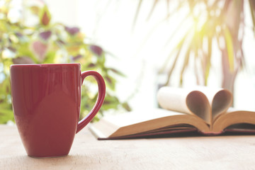 cup and open book on wooden table in front of the window