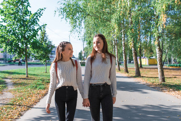 Teenage girls sweaters walking with backpacks behind in summer autumn spring in city, weekends after school. Background trees grass, park. Happy smiling come back from school and college.