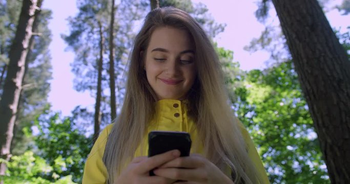 Young Smiling Woman with Mobile Phone in a Forest. Girl in a Yellow Coat in the Woods. Pretty Student Girl using a Cell within tree foliage at a Green Park Path with Natural Sun Light