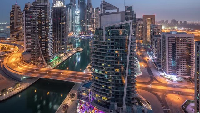 Aerial panoramic view of Dubai Marina residential and office skyscrapers with traffic on a bridge and waterfront night to day transition timelapse before sunrise. Floating boats and yachts