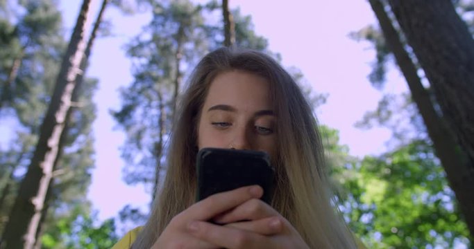  Young Woman with Mobile Phone in a Forest. Girl in a Woodland Glade wearing a Yellow Cagoul. Pretty, blonde Student Girl using a Cell within Tree foliage in a Green Nature Park