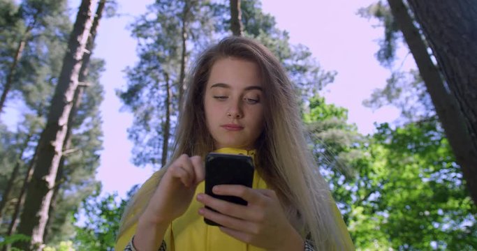  Young Smiling Woman with Mobile Phone in a British Forest. Girl in Woodland Glade wearing a Yellow Rain Coat. Pretty, blonde Student Girl Swiping her Cell within Tree foliage in a Green Nature Park