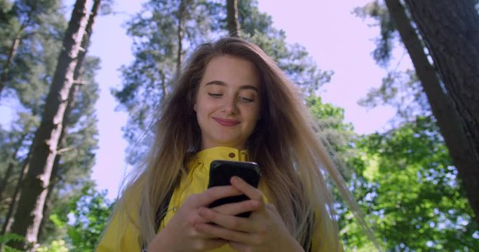  Young Smiling Woman with Mobile Phone in a British Forest. Girl in Woodland Glade wearing a Yellow Rain Coat. Pretty, blonde Student Girl Swiping her Cell within Tree foliage in a Green Natural Park