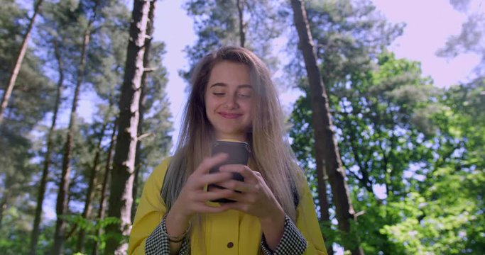 Young Smiling Woman swiping a Mobile Phone in the Forest. Girl in Woodland Glade wearing a Yellow Rain Coat. Happy, blonde Student Girl with Cell Emoji within Trees at a Green Natural Park
