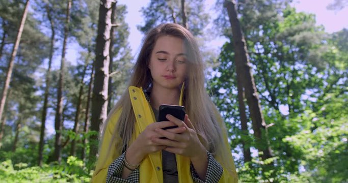 Young Woman texting a Mobile Phone in the Forest. Girl in Wood land Glade wearing a Yellow Rain Coat. Happy, blonde Student Girl with Cell Emoji within Trees at a Green Natural Park
