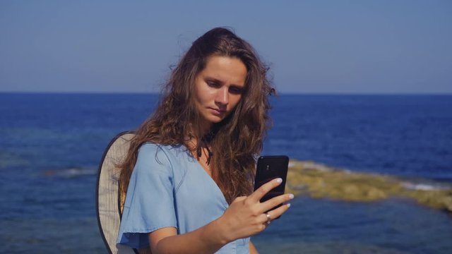 Young brunette woman in a blue dress makes selfie on the beach on a background of blue sea