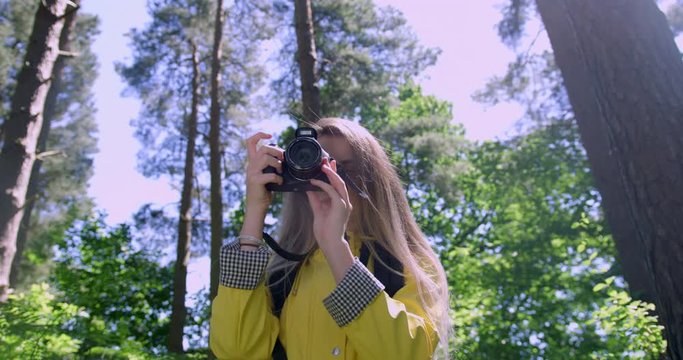 Young Smiling Woman with Camera in the Forest. Female Photographer in a Woodland Glade wearing a Yellow Rain Coat. Happy, Blonde Student Girl within Trees at a Green Festival Park